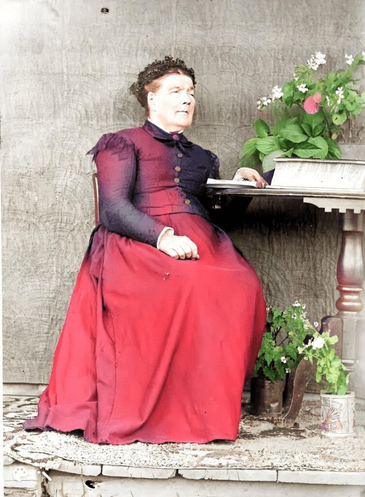 A Colourized picture of a woman sitting with one arm on a small tale.