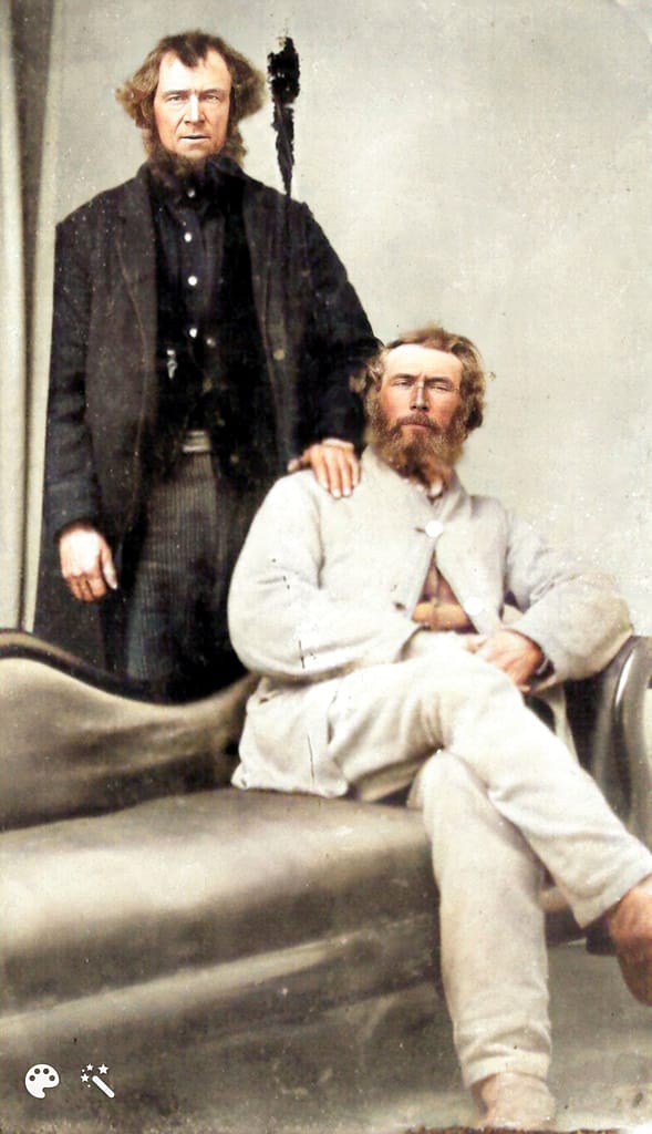 Gentleman in a white suit sitting on a divan with a man behind him with his hand on the sitting man's shoulder.