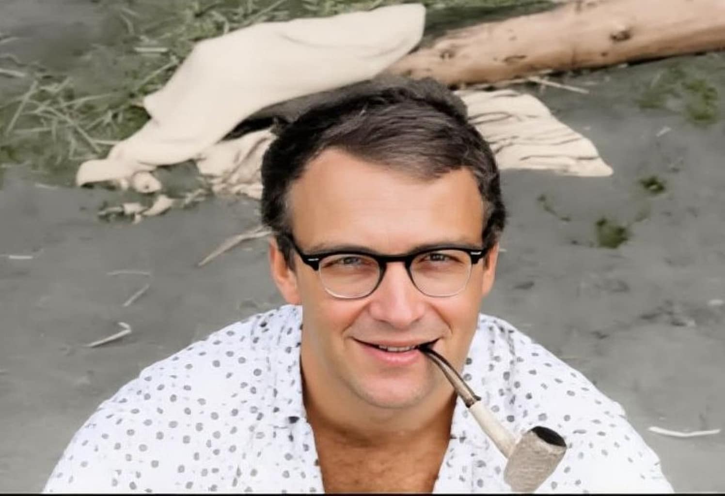 A young man with a white shirt sitting in the sand with a pipe in his mouth.
