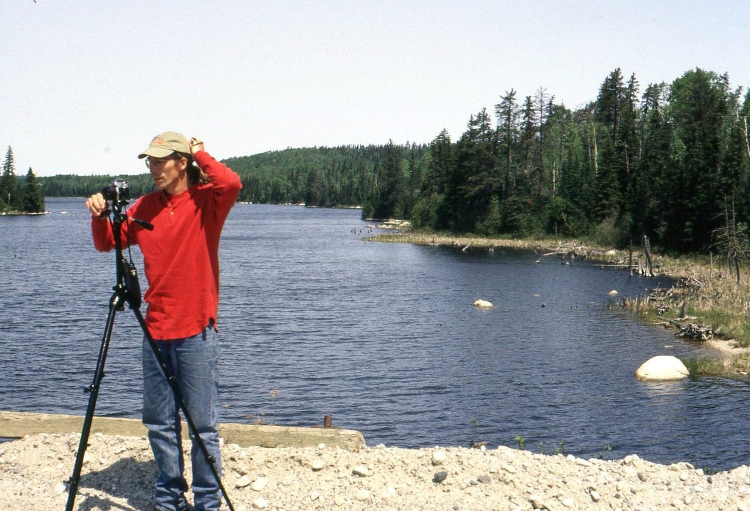 A man by a lake in a red shirt looking in camera on a tripod.