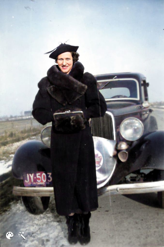 Woman in hat, mufff and black fur collared coat standing in front of her car.