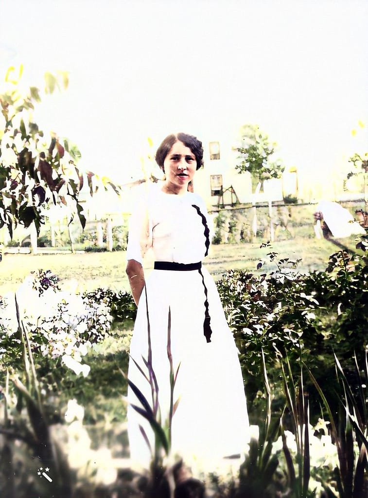 Woman in a white dress with a dark sash posing in a field of flowers.